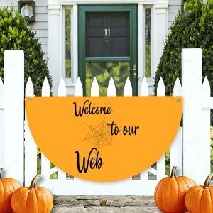 Könnte beinhalten: Ein halbkreisförmiges orangefarbenes Schild mit einer schwarzen Spinnennetzgrafik und dem schwarzen Schriftzug "Welcome to our Web" in Schreibschrift. Das Schild hat zwei Metallösen an den oberen Ecken und ist an einem weißen Lattenzaun befestigt.
