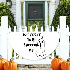 Könnte beinhalten: Ein weißes, halbkreisförmiges Schild mit einer schwarzen Strichzeichnung eines Geistes und dem schwarzen Text "YOU'VE GOT TO BE SHEETING ME!". Das Schild ist an einem weißen Lattenzaun befestigt, mit orangefarbenen Kürbissen im Vordergrund.