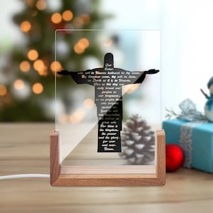 May include: A clear acrylic religious decoration featuring a black silhouette of Jesus with outstretched arms. The text of the Lord's Prayer is printed on the acrylic. The decoration is set in a wooden base and is lit from below.