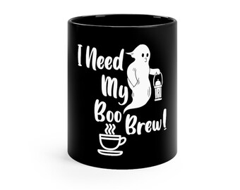 Op de afbeelding: Zwarte keramische mok met de tekst "I Need My Boo Brew!" in het wit. Een witte geest met een lantaarn is ook te zien. Een witte koffiekop met stoom staat onder de tekst. Een Halloween-thema koffiemok.