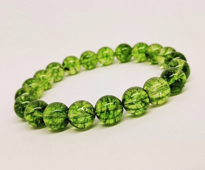 May include: A stretch bracelet composed of round, translucent green beads. Each bead features a distinct internal network of dark green, moss-like inclusions that create a unique, organic pattern throughout the polished, spherical surface of the stones.