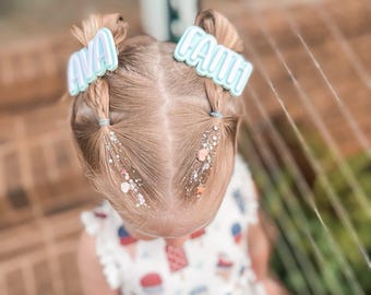 May include: A young child with blonde hair styled in two pigtails, each adorned with a light blue and white hair clip spelling out "AVA" and "FAITH". The child wears a white dress with a red, white, and blue ice cream cone pattern.