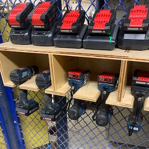 May include: A wooden storage rack with multiple compartments holding several cordless power drills and their corresponding battery chargers. The drills are blue and black, while the battery chargers are black with red accents.