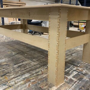 May include: A large rectangular table constructed from light-colored plywood. The table features a flat top surface supported by four thick legs, each assembled with visible interlocking finger joints along the vertical edges and horizontal support beams.
