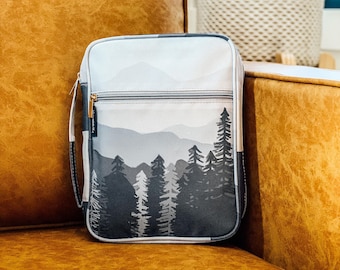 Può includere: Borsa a tracolla grigia e bianca con un design di montagna e foresta. La borsa ha una chiusura con cerniera dorata e un cinturino nero. La borsa è appoggiata su un divano in pelle marrone.