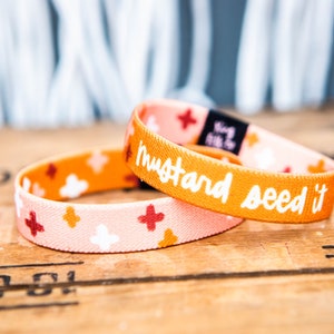 May include: Two colorful elastic bracelets with a mustard yellow background and white polka dots. The top bracelet has the text "mustard seed it" in white lettering.