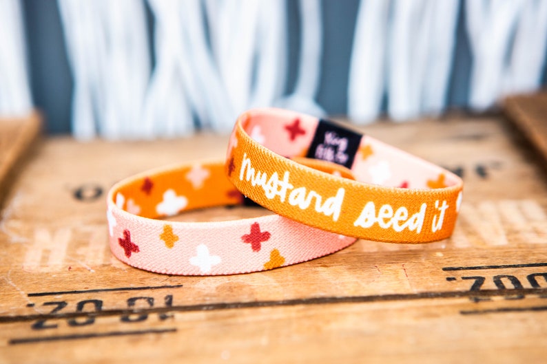 May include: Two colorful elastic bracelets with a mustard yellow background and white polka dots. The top bracelet has the text "mustard seed it" in white lettering.