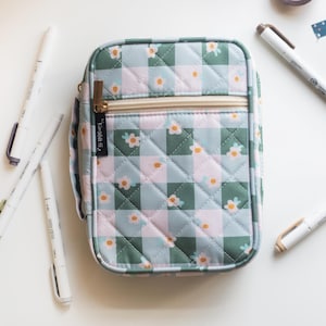May include: A quilted pencil case with a floral pattern in shades of green, pink, and white. The case has a gold zipper and is surrounded by white pens and markers. The case measures approximately 8 inches long and 5 inches wide.