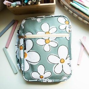 May include: A green zippered pouch with a white daisy pattern. The pouch has a gold zipper and a black label with white text.