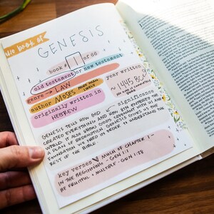 May include: A close-up of a Bible open to the book of Genesis. The page is filled with handwritten notes about the book, including the genre, author, year written, and key verses. The notes are written in colourful markers on a white background.