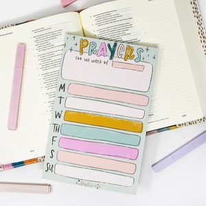 May include: An open book with a colorful cover and a notepad that says "Prayers for the week of" with spaces for each day of the week. There are three light purple highlighters. The notepad has pastel colored rectangles for each day of the week.