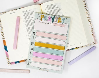 May include: An open book with a colorful cover and a notepad that says "Prayers for the week of" with spaces for each day of the week. There are three light purple highlighters. The notepad has pastel colored rectangles for each day of the week.