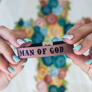 May include: A pink fabric wristband with black text that reads "MAN OF GOD".