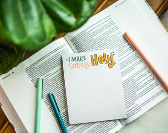 May include: A white notepad with the words "MAKE today HOLY" written in colorful lettering. The notepad is on top of an open Bible with two green and blue pens.