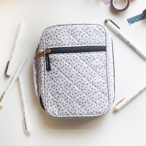 May include: A white and gray polka-dot pencil case with a black zipper and gold zipper pull. The case is surrounded by white pens, washi tape, and a small wooden box. The case has a quilted pattern.