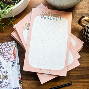 May include: A stack of pink notepads with a white dotted background and the text "Sermon Notes" in gold lettering. The notepads are on a wooden surface with a cup of coffee and a plant in a white pot.