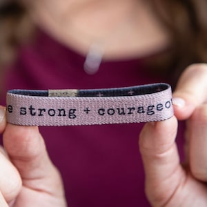 May include: A pink and blue stretchy bracelet with the text "be strong + courageous" printed in black.