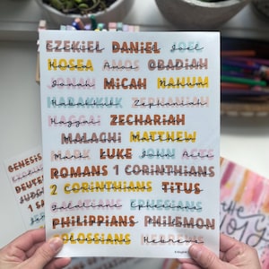 May include: A white sheet of paper with colorful stickers featuring the names of books in the Bible. The stickers are in various colors, including pink, yellow, blue, and brown. The text on the stickers includes "Genesis", "Exodus", "Deuteronomy", "Judges", "1 Samuel", "2 Samuel", "1 Kings", "2 Kings", "Ezekiel", "Daniel", "Joel", "Hosea", "Amos", "Obadiah", "Jonah", "Micah", "Nahum", "Habakkuk", "Zephaniah", "Haggai", "Zechariah", "Malachi", "Matthew", "Mark", "Luke", "John", "Acts", "Romans", "1 Corinthians", "2 Corinthians", "Titus", "Galatians", "Ephesians", "Philippians", "Philemon", "Colossians", "Hebrews".