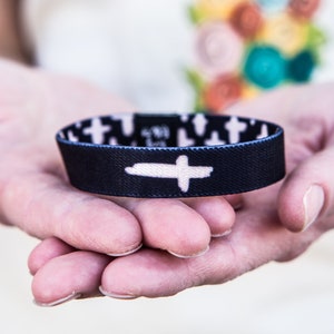 May include: A black and white wristband with a white cross design. The wristband is made of fabric and has a stretchy fit.