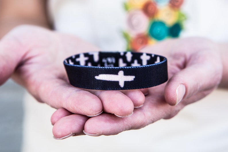 May include: A black and white wristband with a white cross design. The wristband is made of fabric and has a stretchy fit.