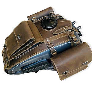May include: Brown leather motorcycle tank bag with two side bags. The tank bag has a large flap with buckles and a small pocket on the front. The side bags have a single buckle closure.