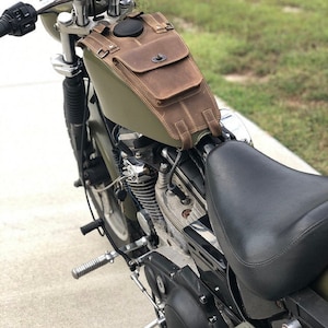May include: A green motorcycle with a brown leather saddlebag attached to the petrol tank. The motorcycle has a black leather seat and a chrome exhaust pipe.