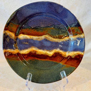 May include: A handmade ceramic plate with a unique, layered design. The plate features bands of blue, orange, and green, with a wavy pattern. The plate is displayed on a clear stand, showcasing its artistic craftsmanship.