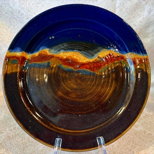 May include: A handmade ceramic plate with a unique design. The plate features a deep blue rim that transitions into a band of orange, red, and brown hues, and a dark brown center. The plate is supported by a clear stand.