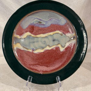 May include: A handmade ceramic plate with a dark green rim and a center featuring red, blue, and yellow abstract designs. The plate is displayed on a clear plastic stand, showcasing its unique, artistic style.