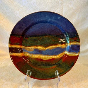 May include: A round ceramic plate with a glossy finish. The plate features a color palette of deep blue, red, green, and gold. The design includes horizontal bands of color, creating a layered effect. The plate is displayed on a clear stand.
