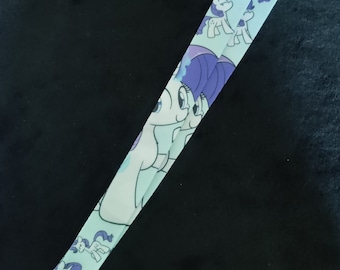 May include: A light blue lanyard with a repeating pattern of cartoon ponies. The ponies are purple and white with blue eyes. The lanyard has a silver metal clip.