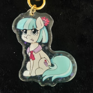 Może przedstawiać: Przezroczysty brelok akrylowy z kreskówkowym kucykiem o jasnoniebieskich włosach, różowej kokardce i jasnoniebieskim ogonie. Kucyk siedzi i ma błyszczące tło. Brelok jest przymocowany do złotego klipsa w kształcie gwiazdy.