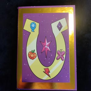 May include: A purple notebook with a gold border and a yellow horseshoe with five colourful gems on it. The gems are a blue teardrop, a purple diamond, a pink star, a red apple, and a pink butterfly.