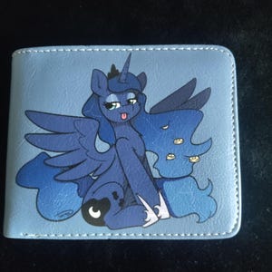 Könnte beinhalten: Eine blaue Lederbörse mit einem aufgedruckten Bild eines blauen und violetten Einhorns mit einer rosafarbenen Mähne und einem gelben Horn. Das Einhorn hat einen blauen Flügel und trägt ein blaues und weißes Kleid.