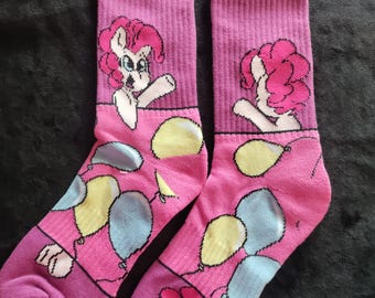 May include: A pair of pink socks featuring a cartoon pony design with pink hair and a white body. The socks have a balloon pattern in yellow, blue, and pink. The socks are made of a soft material.