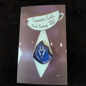 May include: A blue and gold enamel pin featuring a stylized crystal with a blue and white symbol inside. The pin is on a card with the text "Taurson's Cafe Final Fantasy XIV".