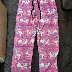 May include: Pink pyjama trousers with a repeating pattern of a pink pony with a blue mane and tail. The trousers have a black drawstring.