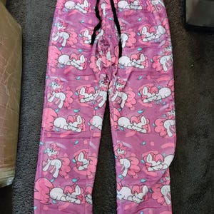 May include: Pink pajama trousers featuring a repeating pattern of a cartoon pony with a pink mane and tail. The trousers have a drawstring waist and are made of a soft, plush material. The background is a lighter shade of pink.