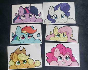 May include: Set of six cartoon pony stickers. Each sticker features a different pony character with a unique color scheme and design. The ponies are shown peeking over a surface, with their faces and upper bodies visible. The stickers are on a white background.