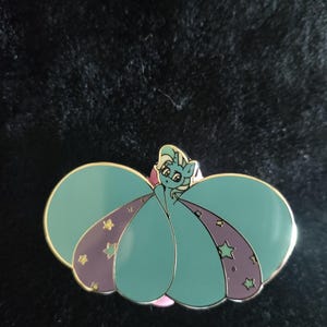 Könnte beinhalten: Ein tealfarbener Emaille-Pin mit einem Cartoon-Pony mit fließendem Kleid. Das Kleid hat lila Akzente mit goldenen Sternen. Der Pin hat einen goldenen Rand und eine rosa Basis. Das Pony hat eine helltürkise Mähne und ein weißes Gesicht.