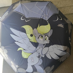 May include: A dark blue umbrella featuring a cartoon pegasus with yellow hair and large wings. The umbrella has a matching carrying case. The design includes clouds and lightning bolts.