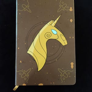 Puede incluir: Una libreta marrón con un diseño de cabeza de unicornio dorado. El unicornio tiene ojos azules y un cuerno dorado.