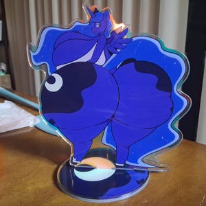 May include: A clear acrylic standee featuring a stylized purple pony character with a dark blue mane and tail, set against a blue background with white star accents. The character is positioned on a circular base with a crescent moon graphic.