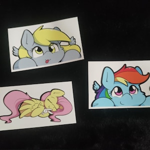 May include: Three cartoon pony stickers. The first sticker features a yellow pony with blue eyes and a light blue mane. The second sticker features a pink pony with yellow wings and a yellow mane. The third sticker features a blue pony with rainbow hair and a yellow wing.
