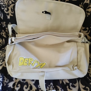 May include: A beige canvas messenger bag with a zipper closure and a yellow graphic that says "DERAYI".