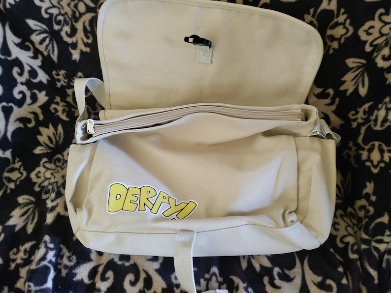 May include: A beige canvas messenger bag with a zipper closure and a yellow graphic that says "DERAYI".
