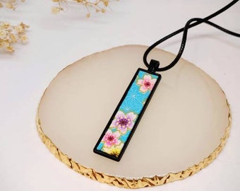 May include: A black necklace with a rectangular pendant featuring a blue and white floral design. The pendant is framed with a black border.