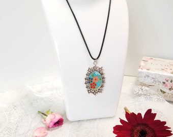 May include: A silver oval pendant necklace with a floral design on a black cord. The pendant features a blue, green, pink, and orange floral pattern with a silver border. The necklace is displayed on a white mannequin.