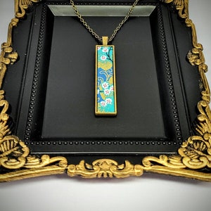 Könnte beinhalten: Eine goldene Kettenkette mit einem rechteckigen Anhänger mit einem blauen und grünen Blumenmuster. Der Anhänger ist in Goldmetall gefasst und hängt an einer goldenen Kette.