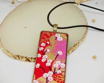 May include: A gold-framed pendant necklace with a red, pink, and gold fabric insert featuring a floral pattern. The fabric is decorated with white flowers and gold accents.